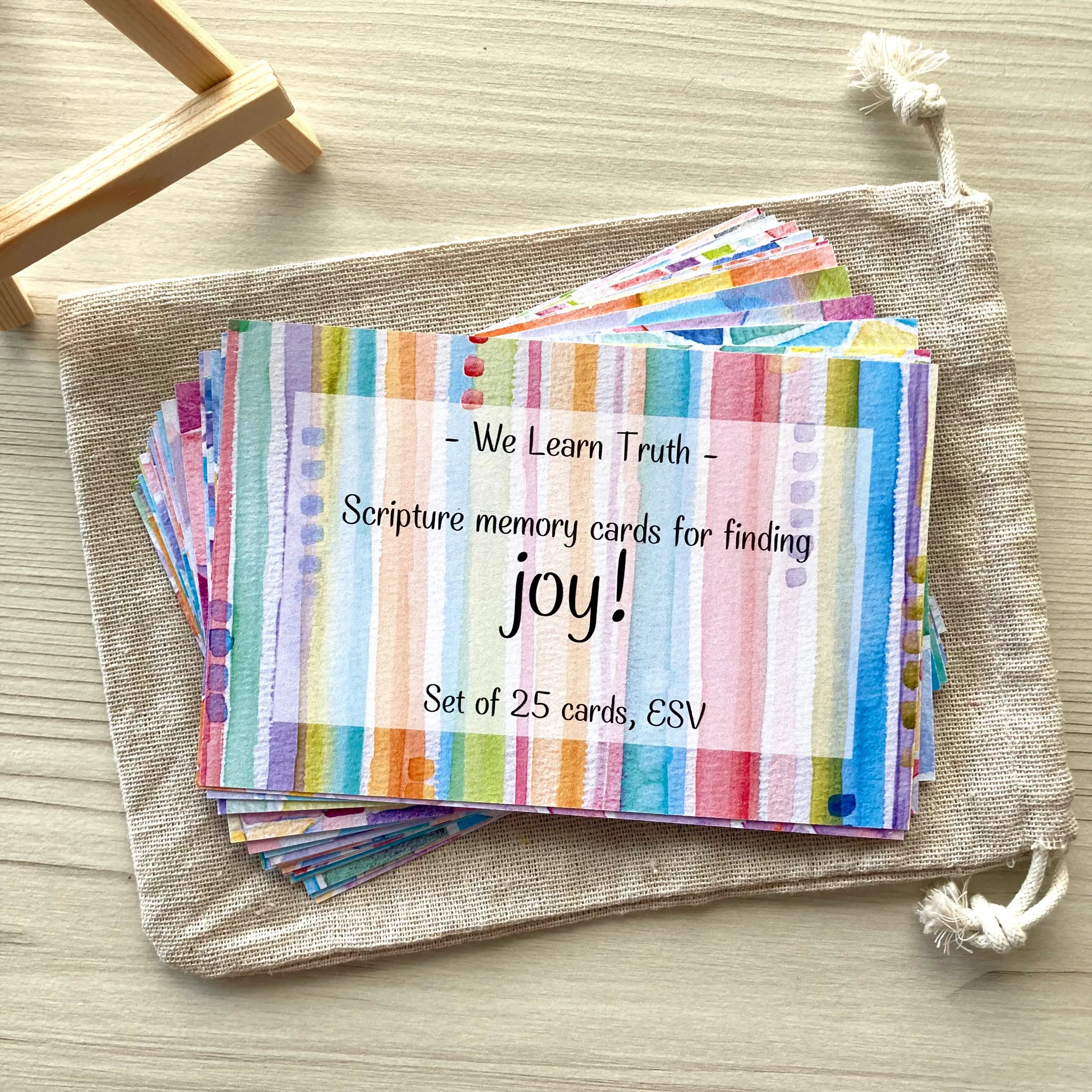 Set of 26 scripture memory cards with watercolor designs in a burlap pouch on a wooden surface. Top card reads 'We Learn Truth,' offering peace and comfort in anxiety, with a plant pot and pink crystal nearby.