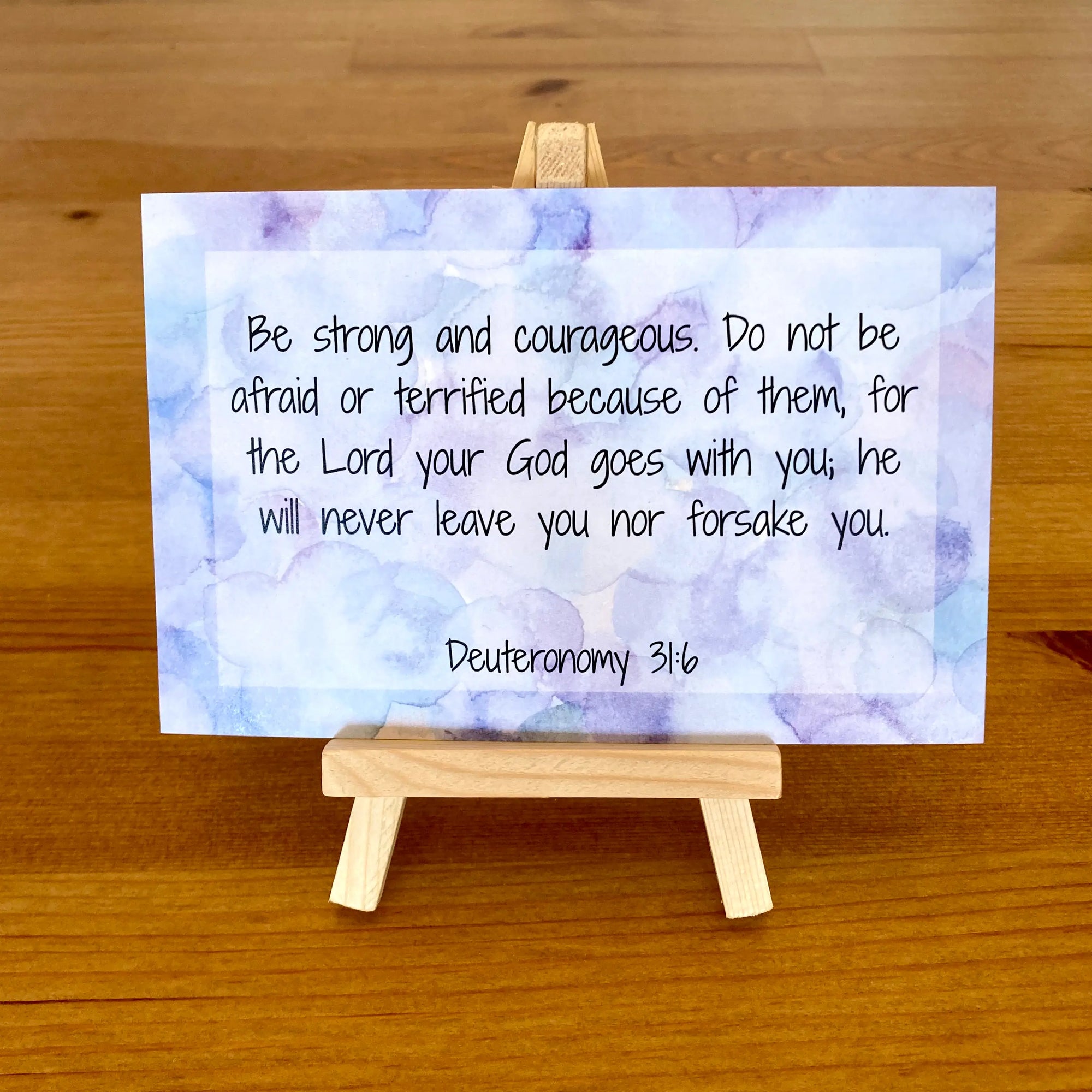Card with Deuteronomy 31:6 text on a colorful geometric background, held by a small wooden easel.