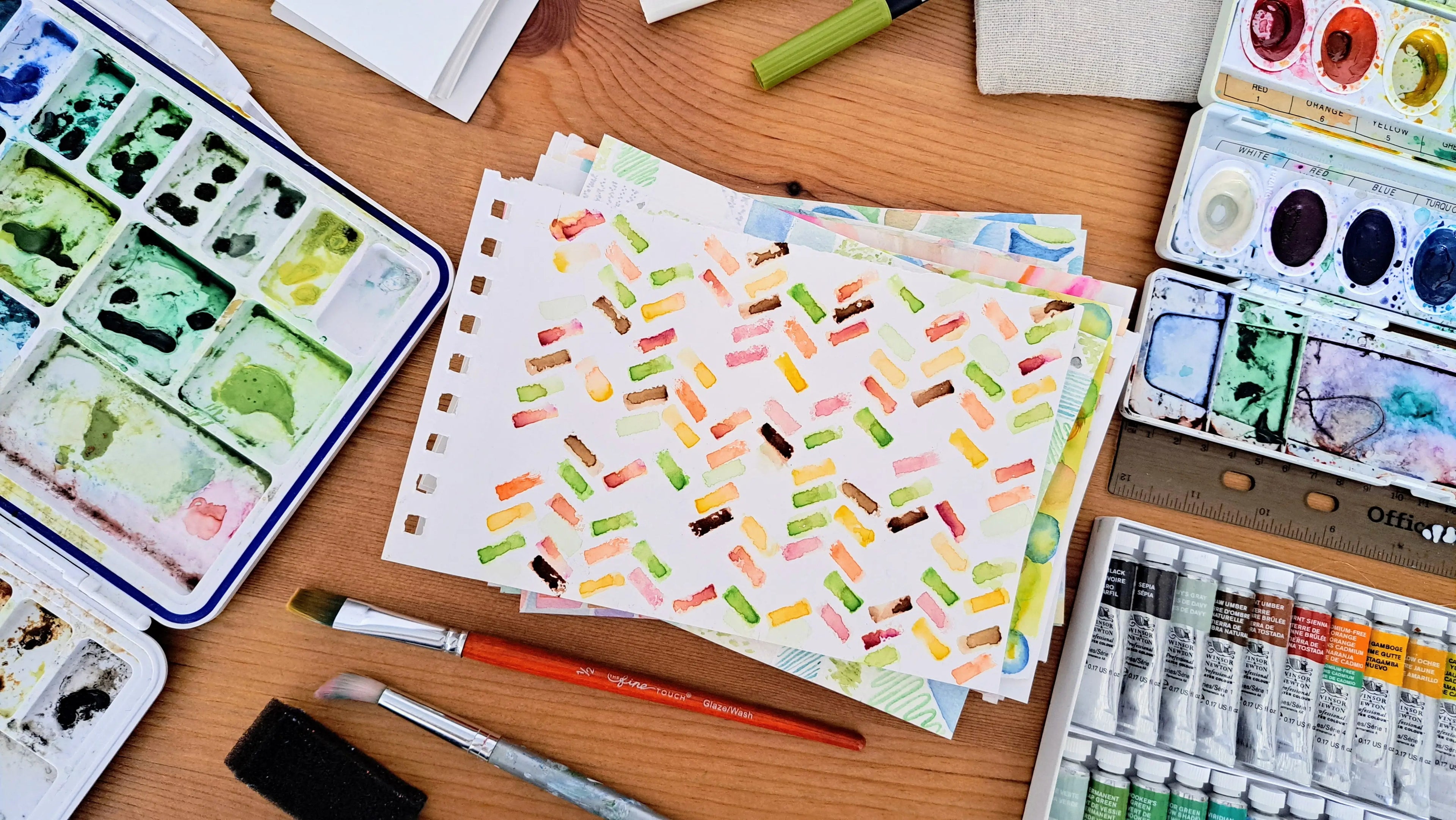 Art supplies including watercolor paints, brushes, and handpainted patterns on a wooden table.