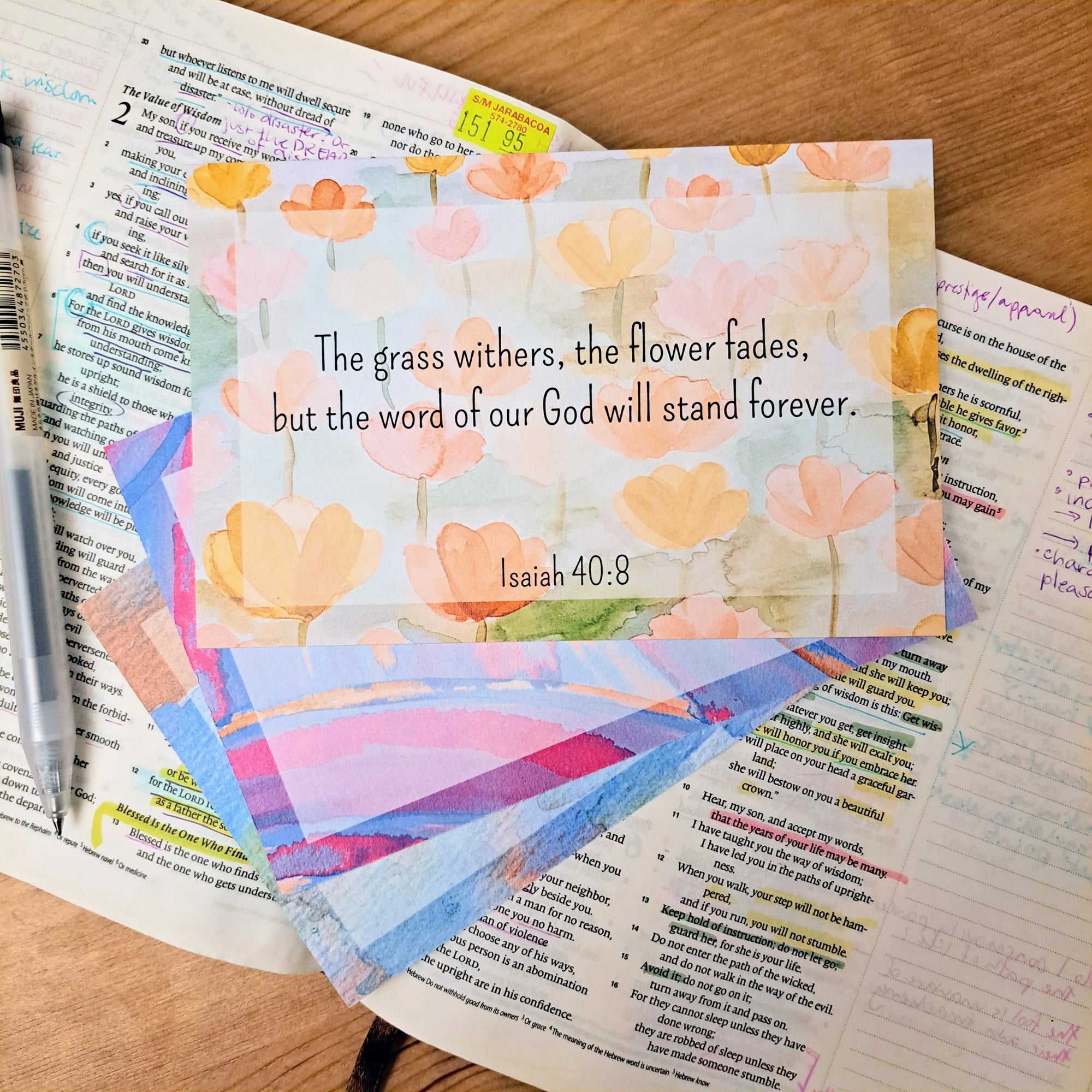 Inspirational card with a floral watercolor background featuring the text "The grass withers, the flower fades, but the word of our God will stand forever. Isaiah 40:8" placed on top of an open bible with handwritten notes and a pen, surrounded by other colorful cards