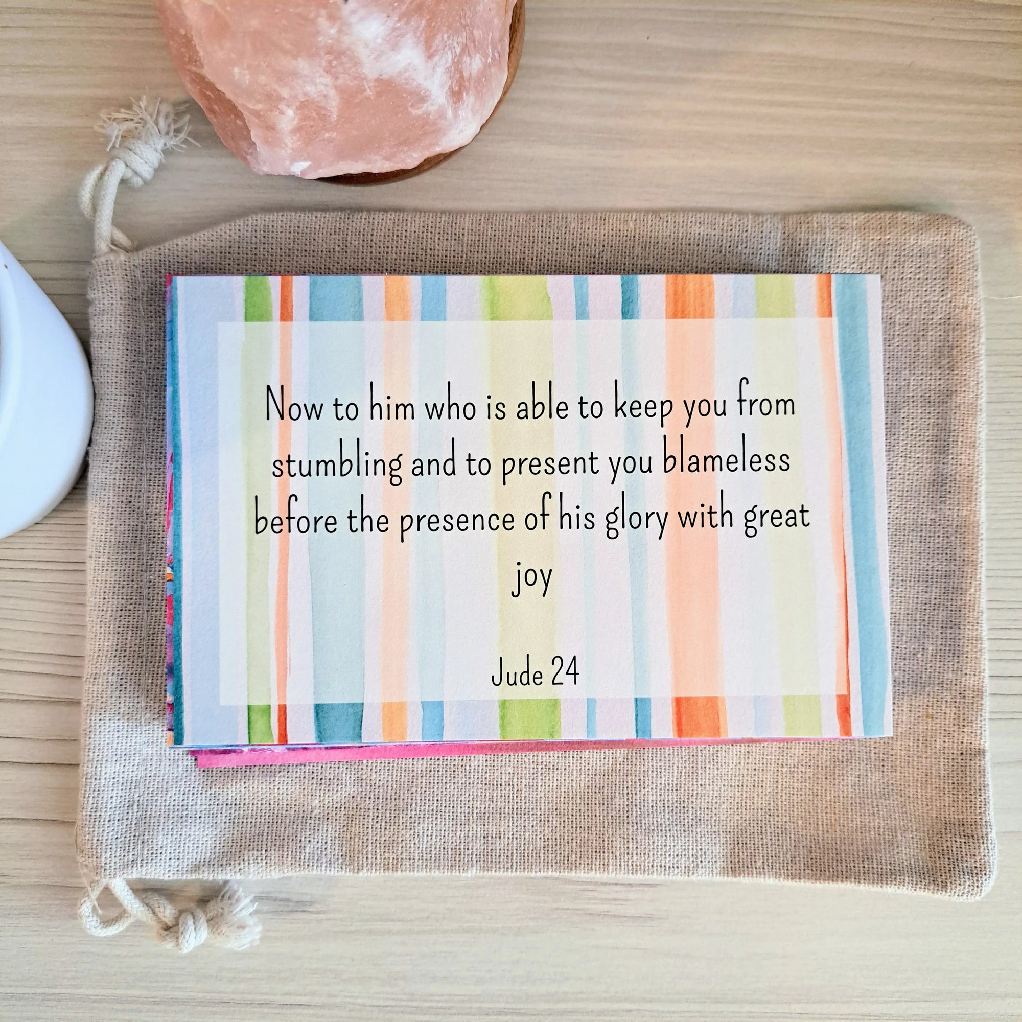 Memorization card with a colorful striped background featuring the text "Now to him who is able to keep you from stumbling and to present you blameless before the presence of his glory with great joy, Jude 24" placed on a canvas pouch with a pink crystal and white object nearby