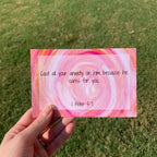 Hand holding a pink and orange spiral card against a grassy background. The card lists 1 Peter 5:7, "cast you anxiety on him because he cares for you."