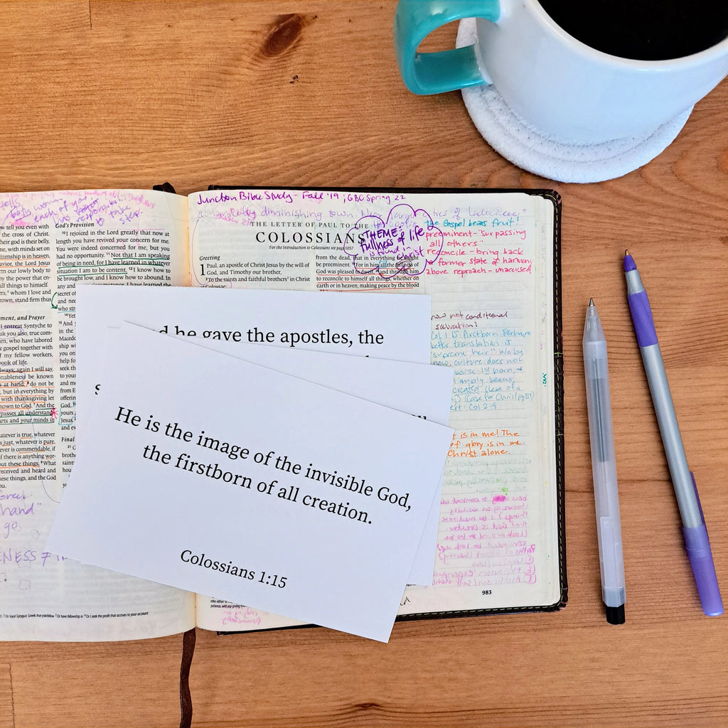 A white scripture memory card with Colossians 1:15 ("He is the image of the invisible God, the firstborn of all creation.") placed on an open, annotated Bible with colorful notes, next to a teal mug and pens on a wooden surface