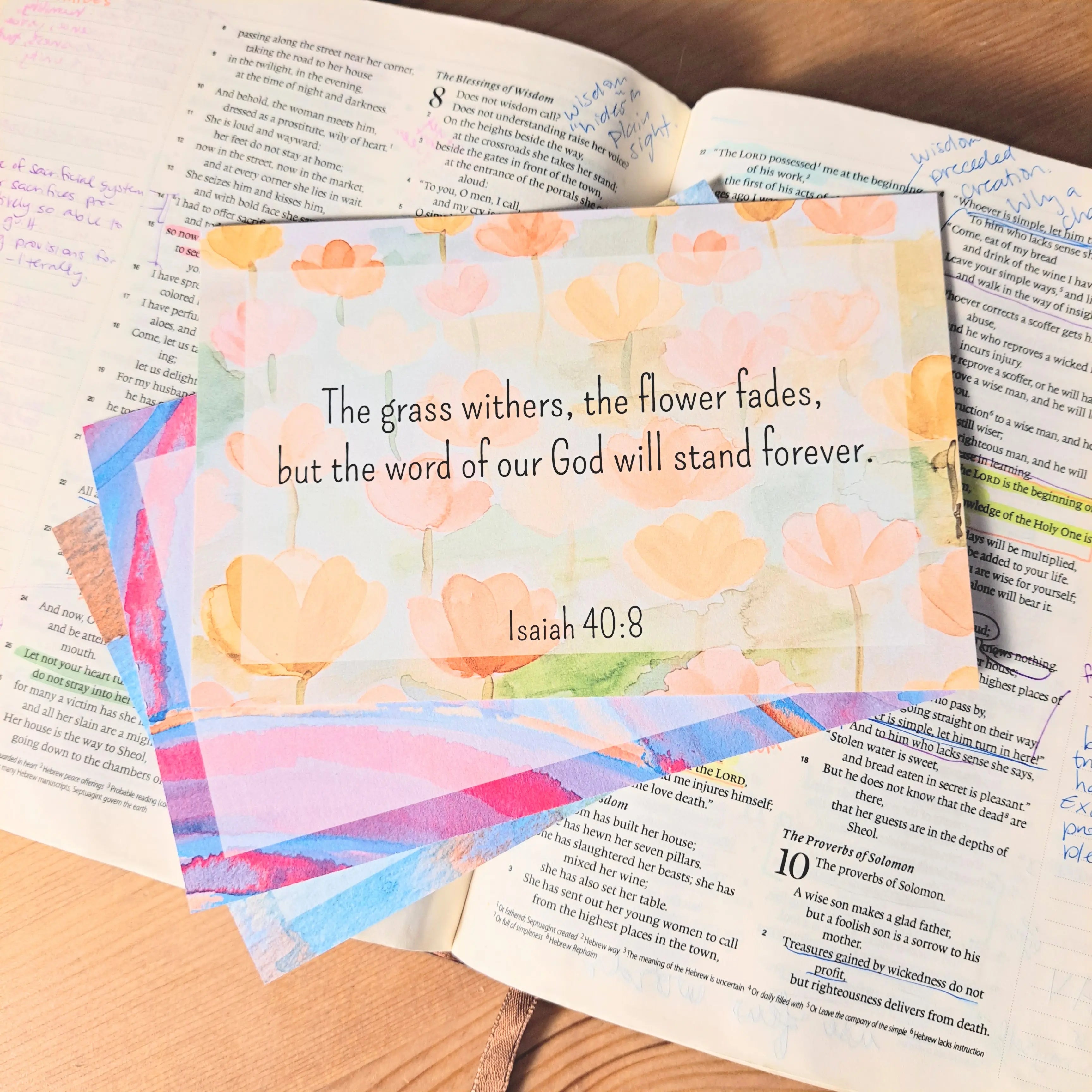 A colorful card with a floral design displays Isaiah 40:8: "The grass withers, the flower fades, but the word of our God will stand forever." It rests on open books on a wooden surface.