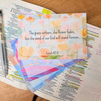 Inspirational card with a floral watercolor background featuring the text "The grass withers, the flower fades, but the word of our God will stand forever. Isaiah 40:8" placed on top of an open bible with handwritten notes and a pen, surrounded by other colorful cards