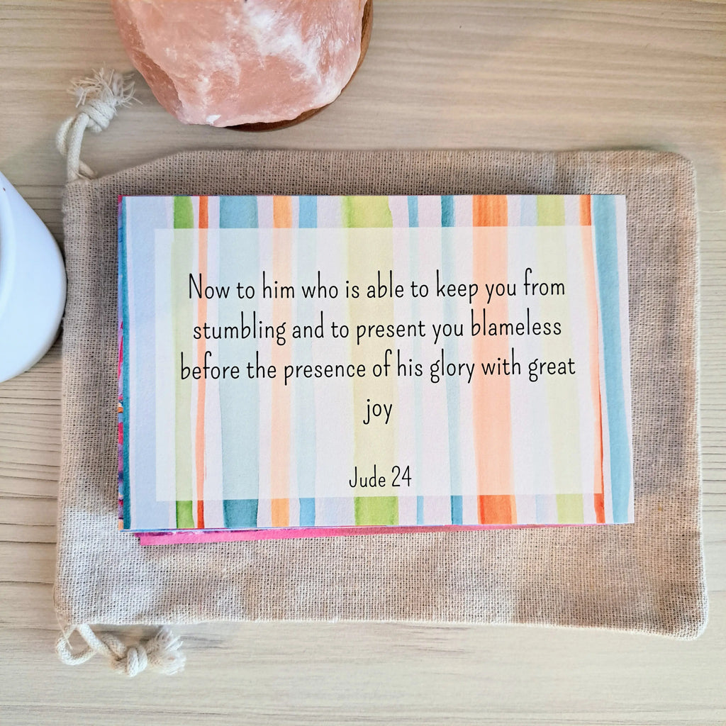 Memorization card with a colorful striped background featuring the text "Now to him who is able to keep you from stumbling and to present you blameless before the presence of his glory with great joy, Jude 24" placed on a canvas pouch with a pink crystal and white object nearby