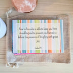 Memorization card with a colorful striped background featuring the text "Now to him who is able to keep you from stumbling and to present you blameless before the presence of his glory with great joy, Jude 24" placed on a canvas pouch with a pink crystal and white object nearby