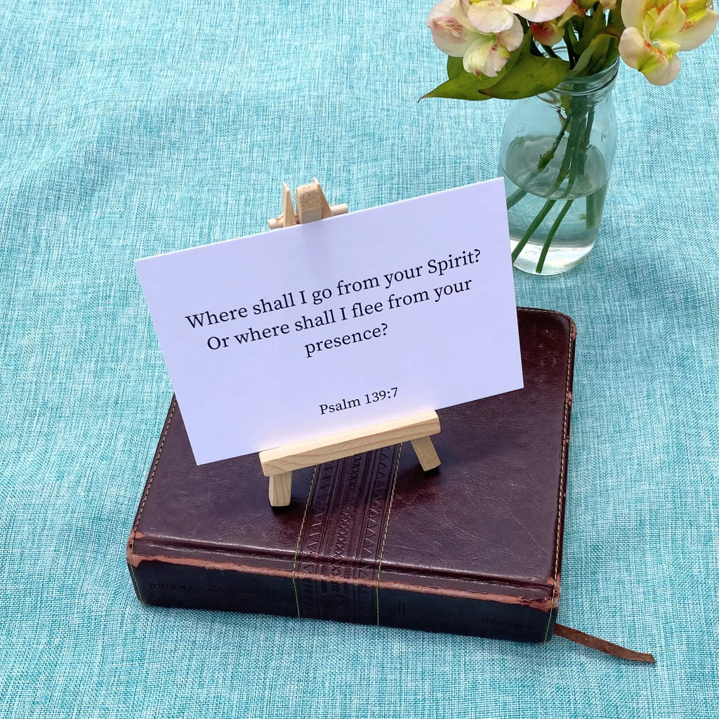 A white card with Psalm 139:7 ("Where shall I go from your Spirit? Or where shall I flee from your presence?") on a wooden stand atop a brown Bible, with a vase of flowers on a teal fabric background