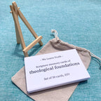 A stack of white scripture memory cards labeled "We Learn Truth - Scripture memory cards of theological foundations, Set of 50 cards, ESV" on a teal fabric, with a canvas pouch and wooden stand nearby